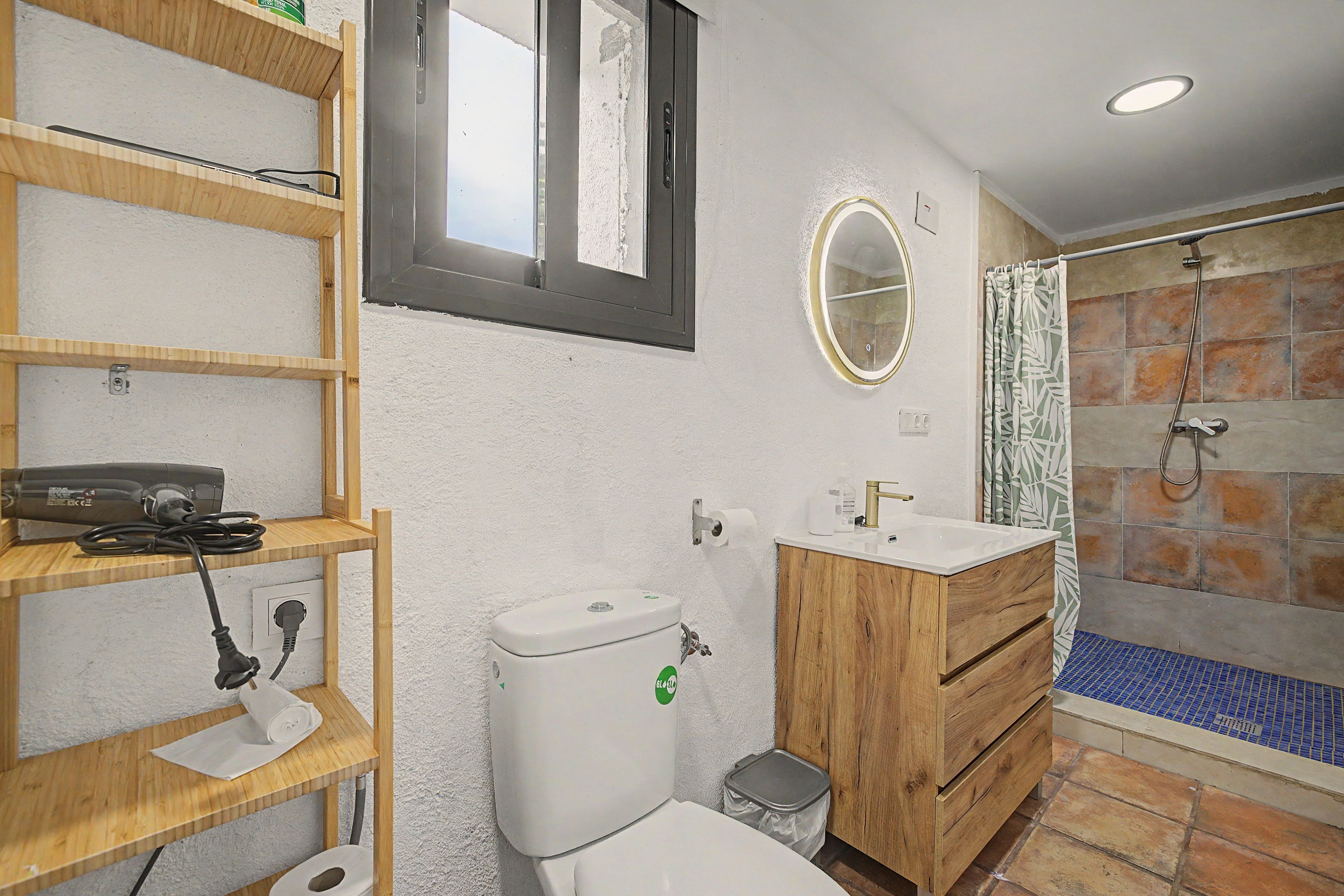 A bathroom with a toilet, a sink, a shower, and a wooden shelf with a hairdryer.