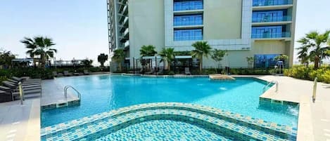 A swimming pool with tiled edges, surrounded by lounge chairs and palm trees, with a tall building in the background.