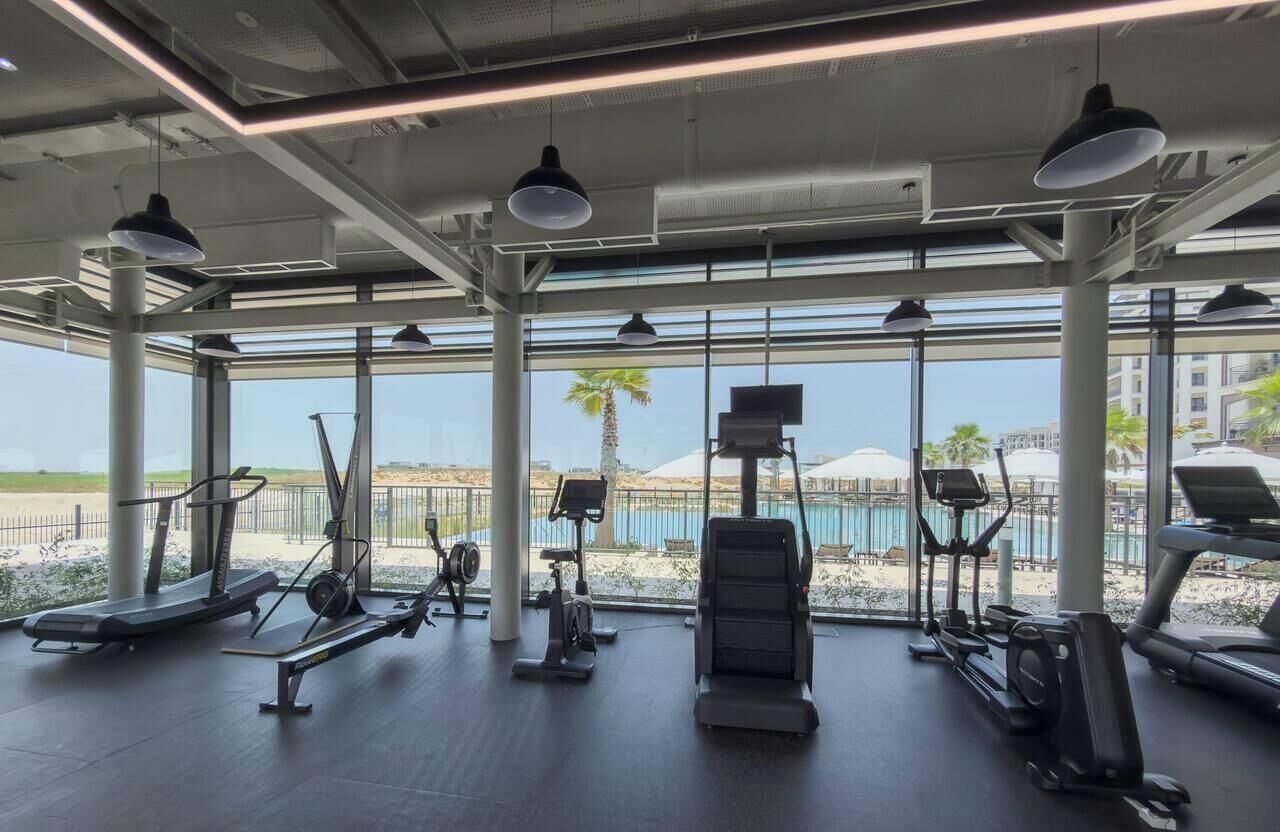 Un gimnasio con equipos de ejercicio variados, grandes ventanales y vistas a una piscina y palmeras.