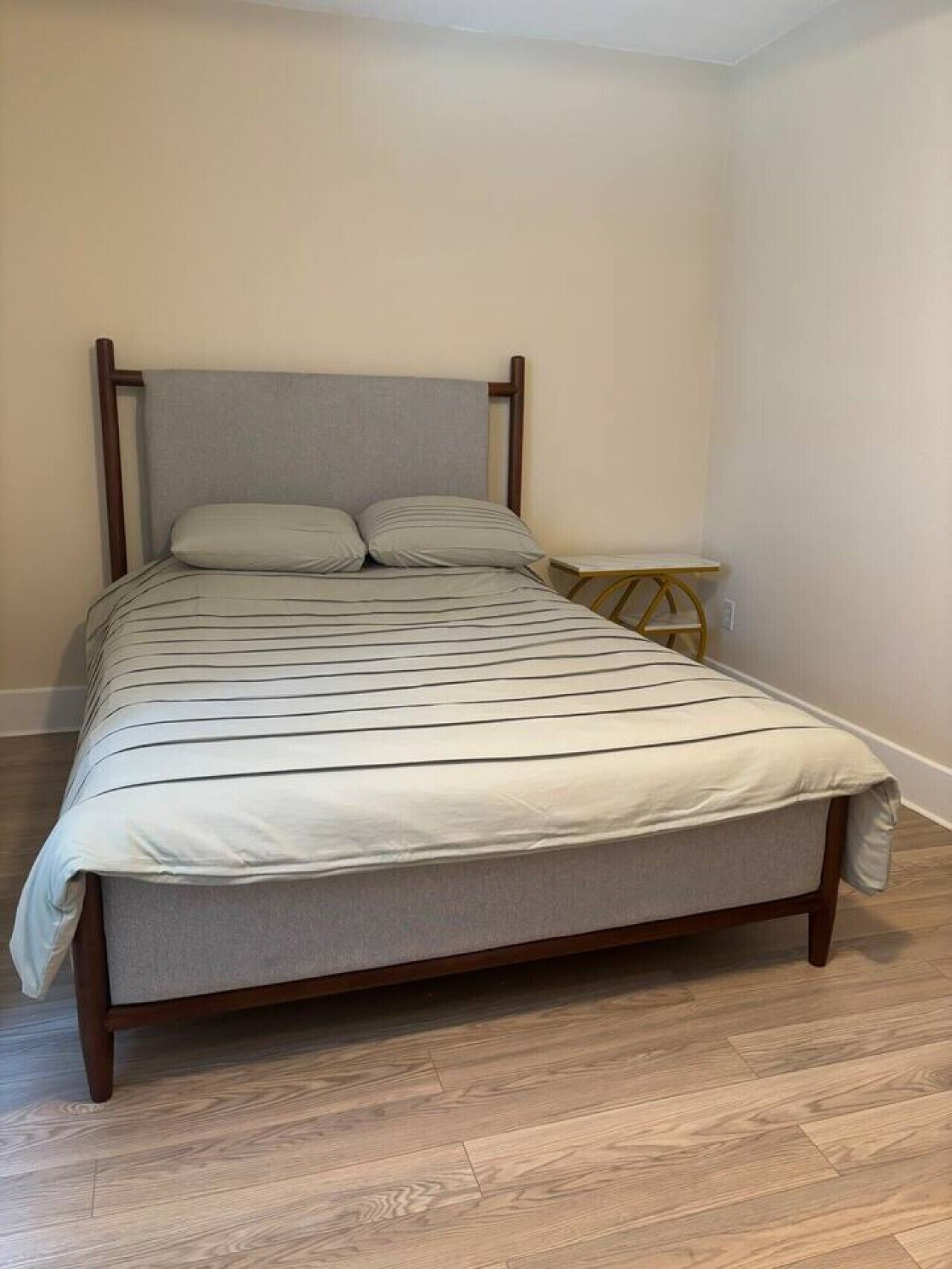 Un lit avec un cadre en bois et une tête de lit grise, une table de chevet et un plancher en bois.