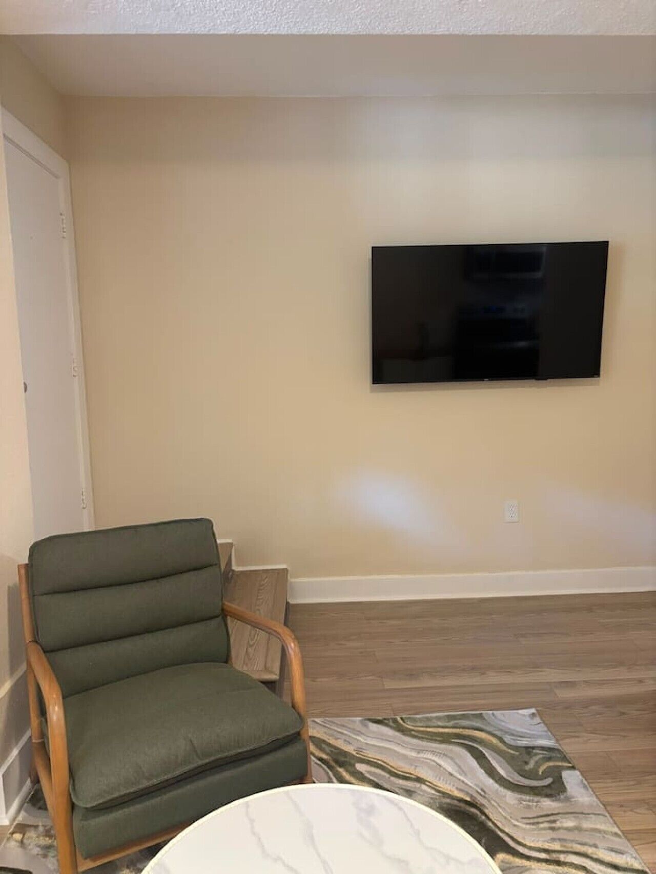 Un salon avec un téléviseur à écran plat fixé au mur, un fauteuil recouvert d’un tissu vert et un tapis à motifs sur le plancher en bois.