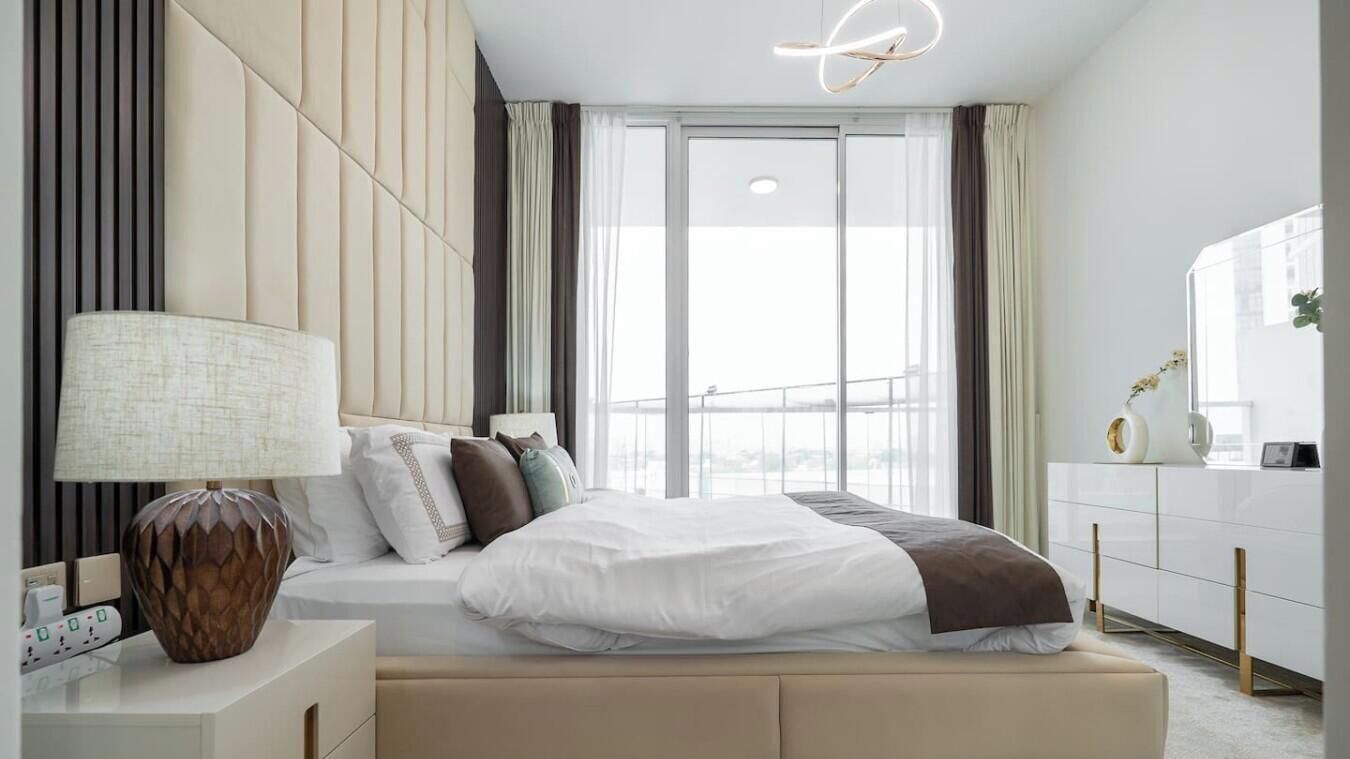 Ein modernes Hotelzimmer mit einem großen Bett, einem Nachttisch mit Lampe, Blick auf die Stadt durch große Fenster und einer weißen Kommode.