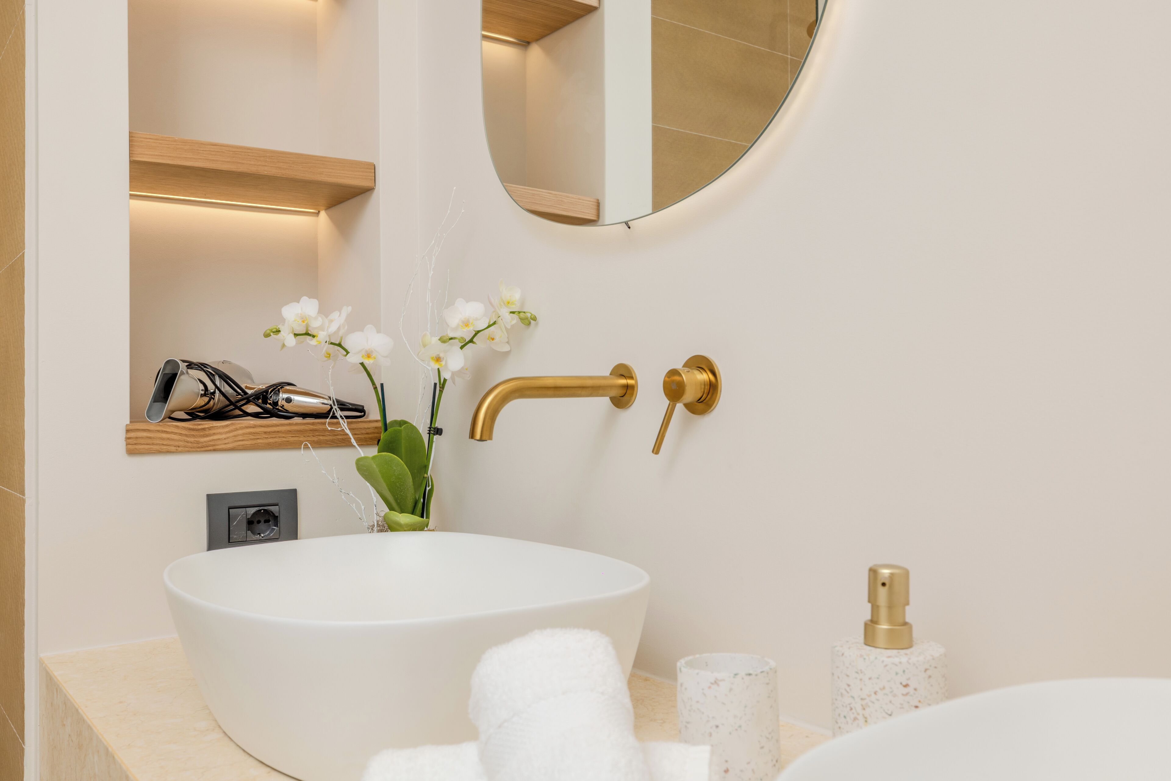 Un baño moderno con un lavamanos ovalado grande, un grifo dorado, un espejo y un estante pequeño con un secador de pelo y orquídeas.