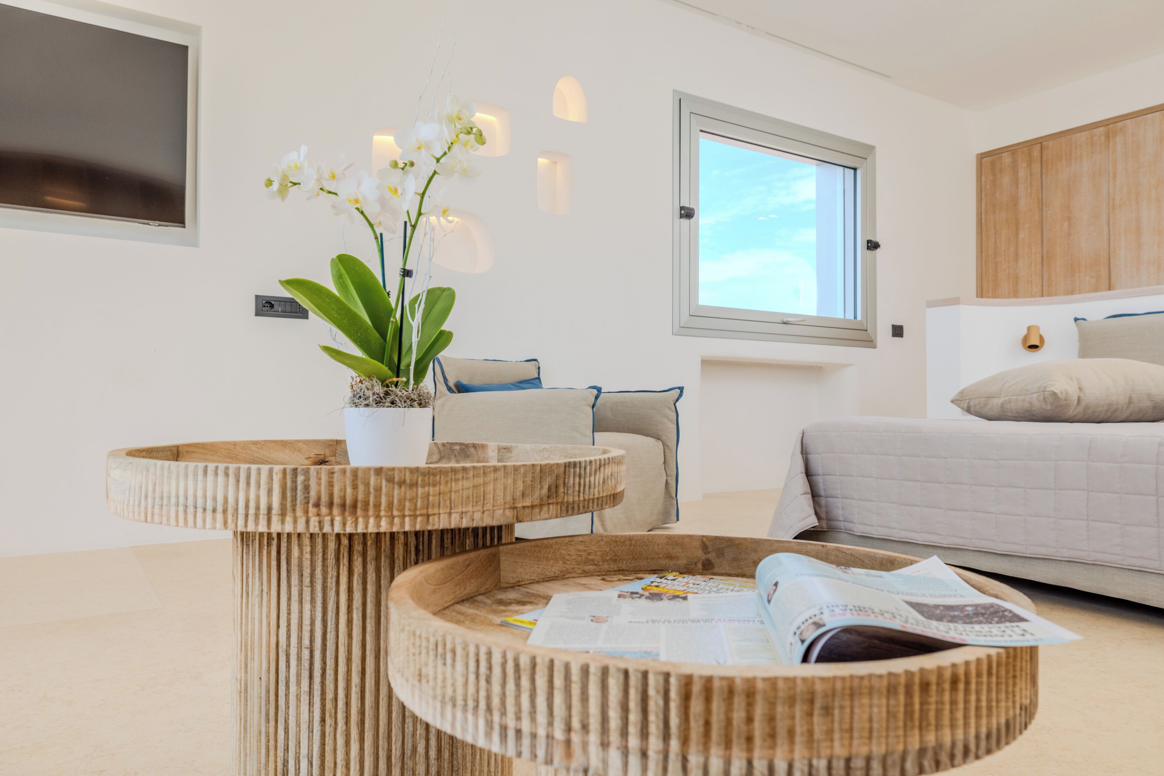 A modern bedroom with a bed, a window, a potted orchid, and a round wooden table with a magazine.