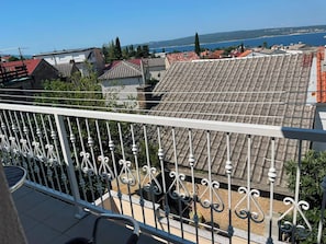 Ein Balkon mit weißer Brüstung, Blick auf terrakottafarbene Dächer und ein entferntes Gewässer.