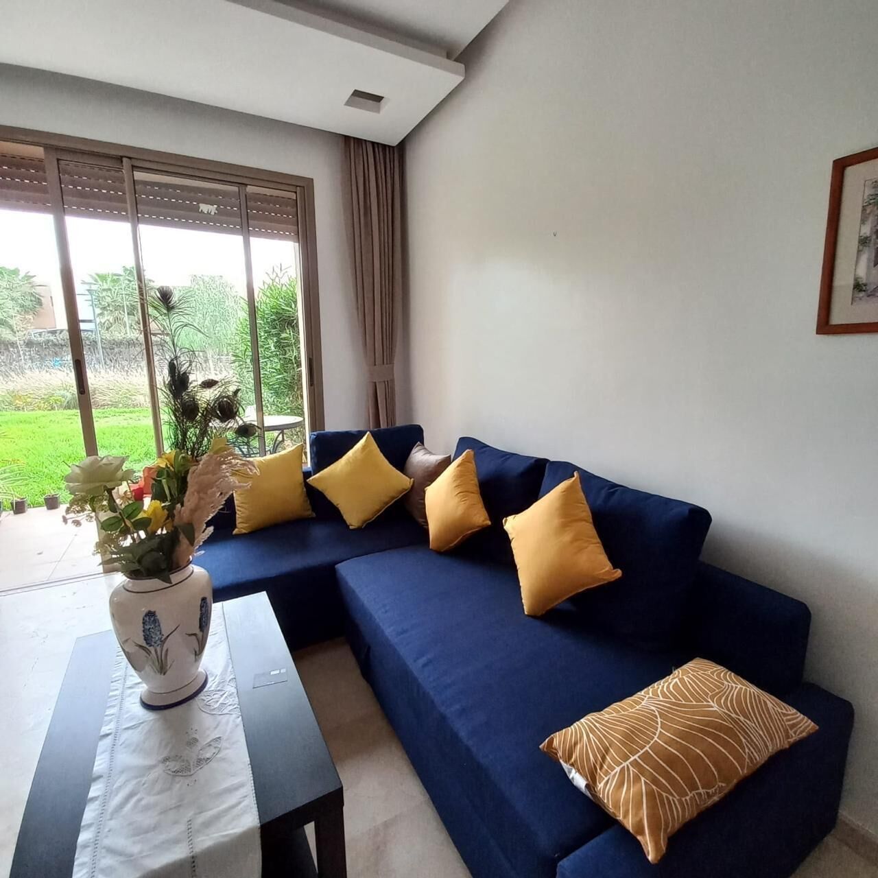 A modern living room with a blue sofa, yellow cushions, a vase with flowers, and a view of greenery outside.