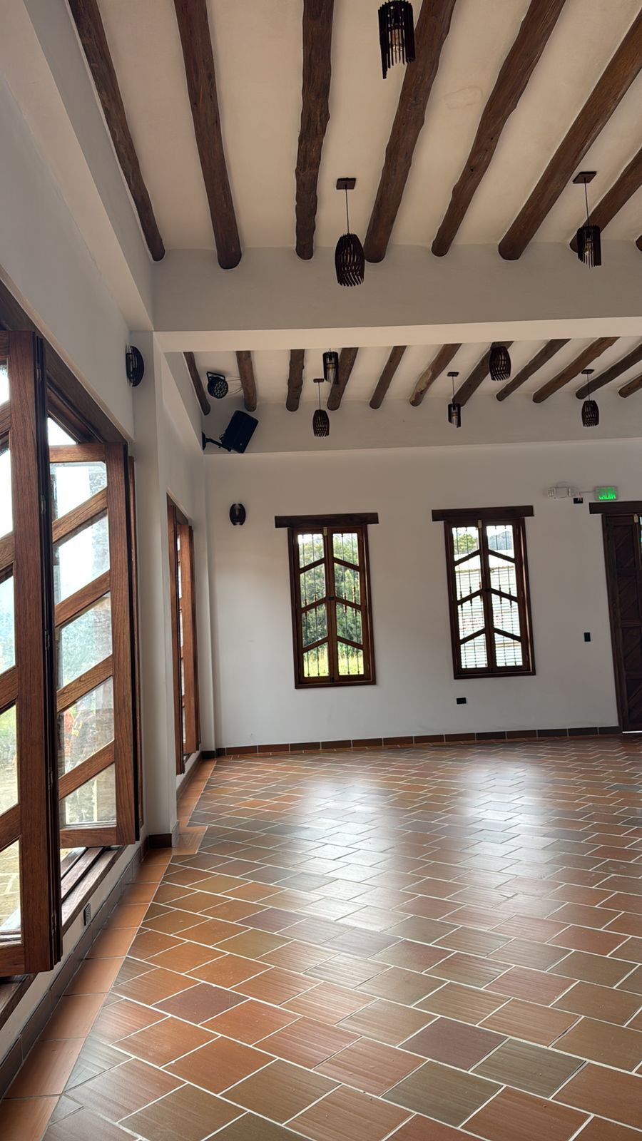 A spacious room with a tiled floor, wooden beams, and large windows.