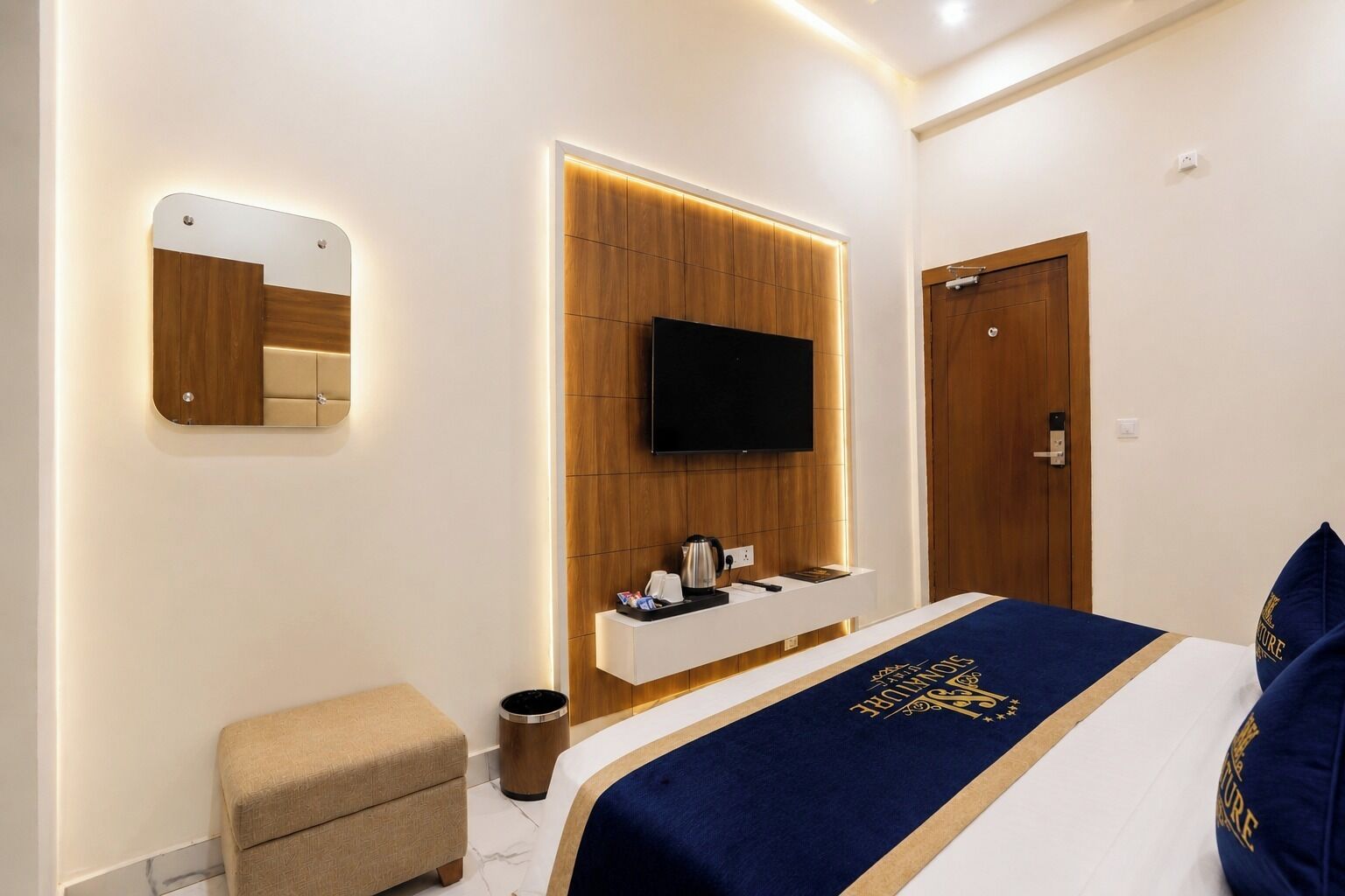 Habitación de hotel moderna con cama, televisor montado en una pared de paneles de madera, mesita de noche con lámpara y un pequeño taburete.