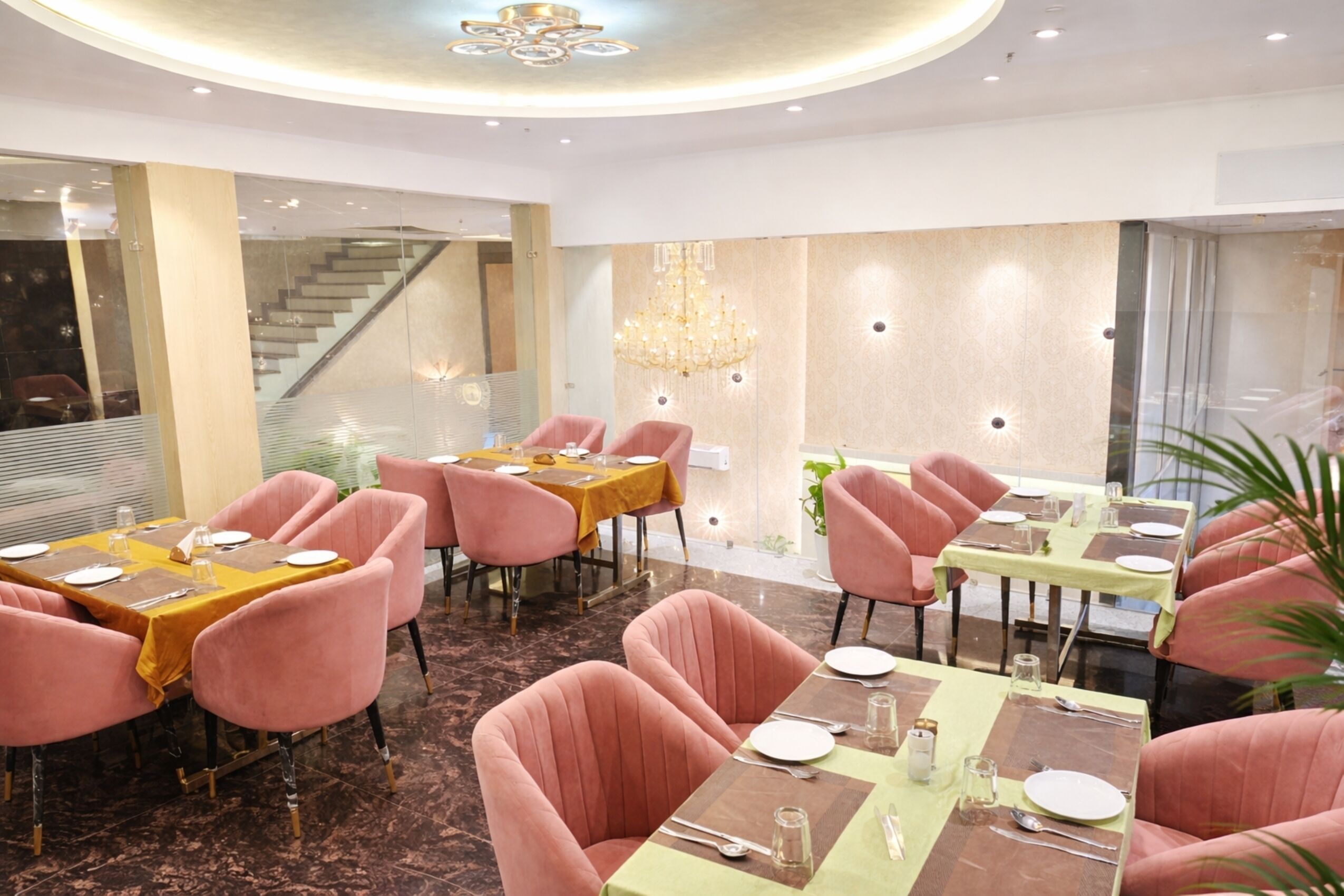 A modern dining area with pink chairs and tables, a chandelier, and a staircase in the background.