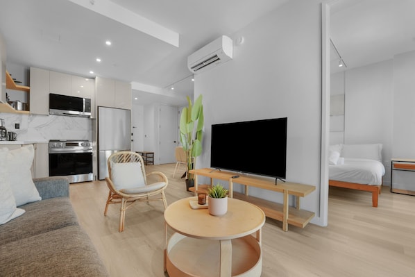 Una sala moderna con un televisor de pantalla plana, una mesa de centro redonda de madera, una silla de mimbre, un sofá y una zona de cocina con microondas y estufa.