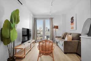 Una sala de estar moderna con un sofá, una silla de mimbre, un televisor y un ventanal con vistas a la ciudad.