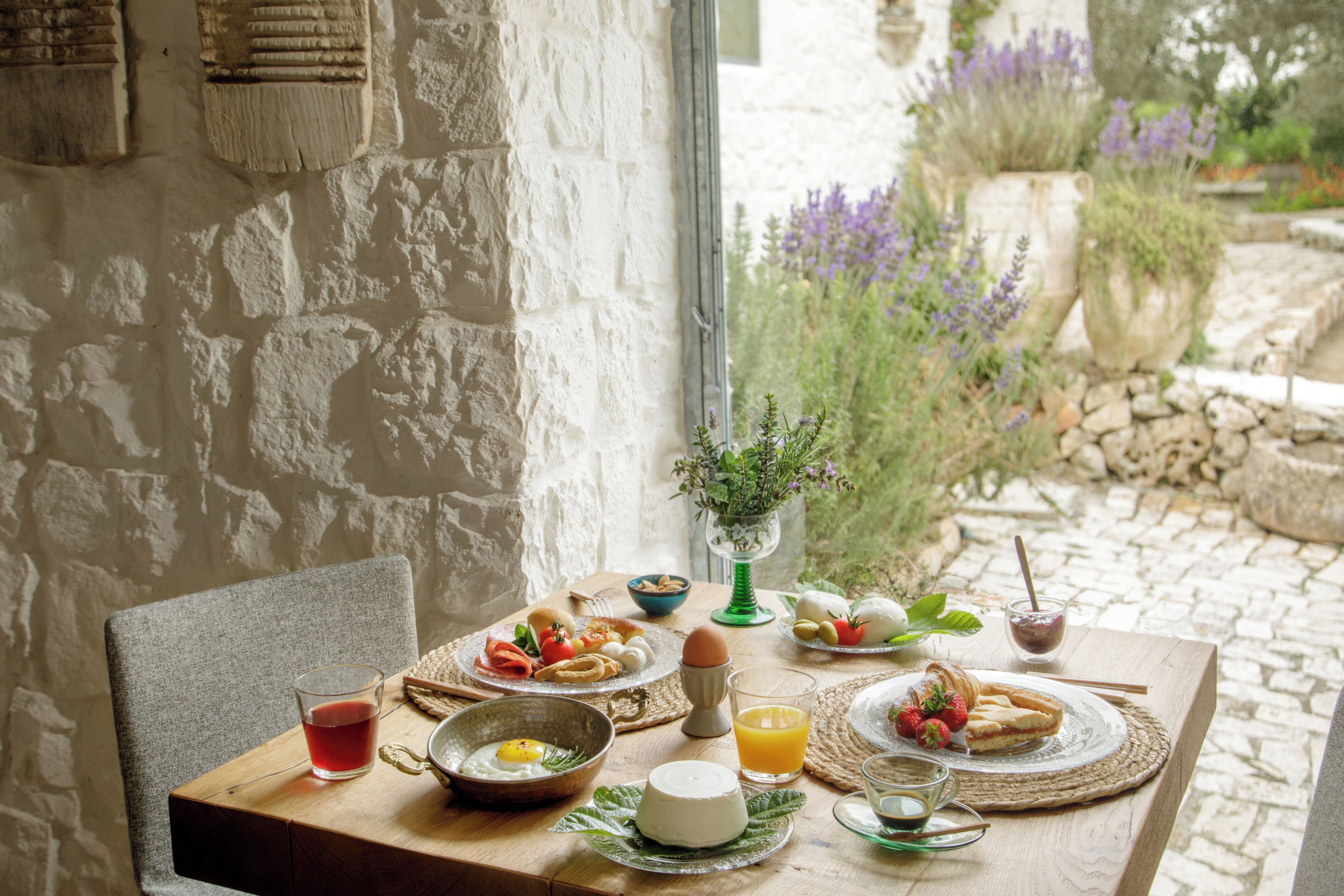 Mesa dispuesta con alimentos para el desayuno, una silla y vista a un jardín con plantas y un muro de piedra.