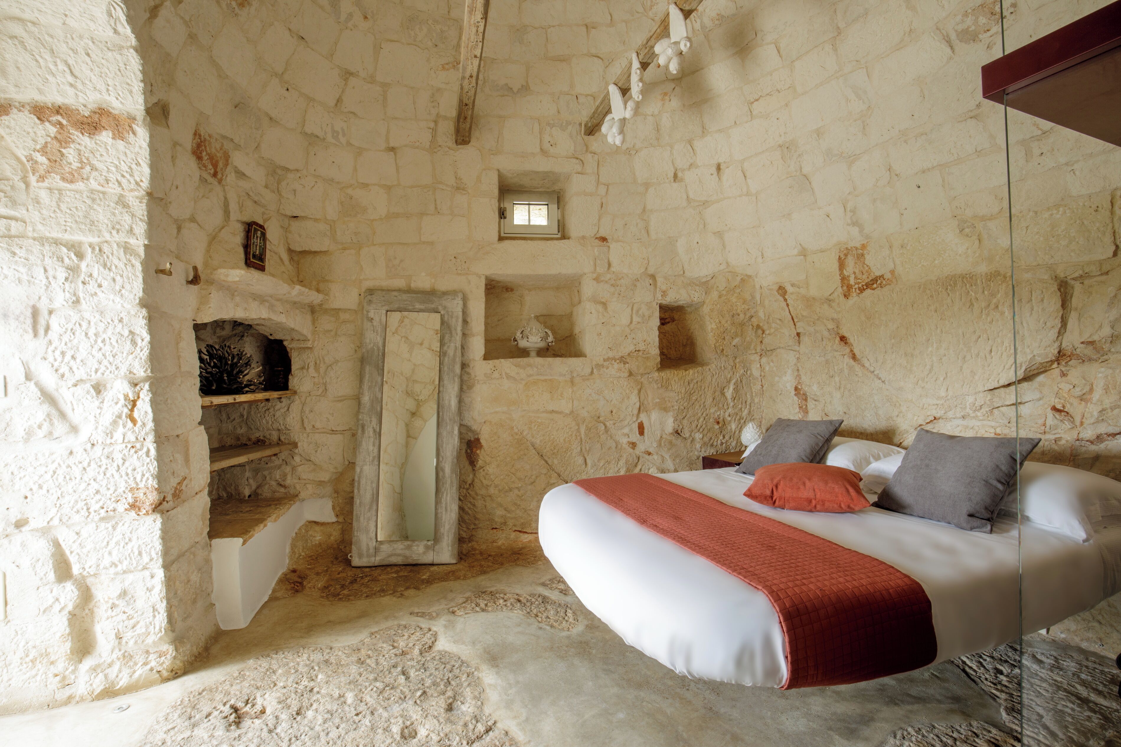 Une pièce aux murs de pierre, avec un lit, une petite fenêtre et une étagère ornée d’objets décoratifs.