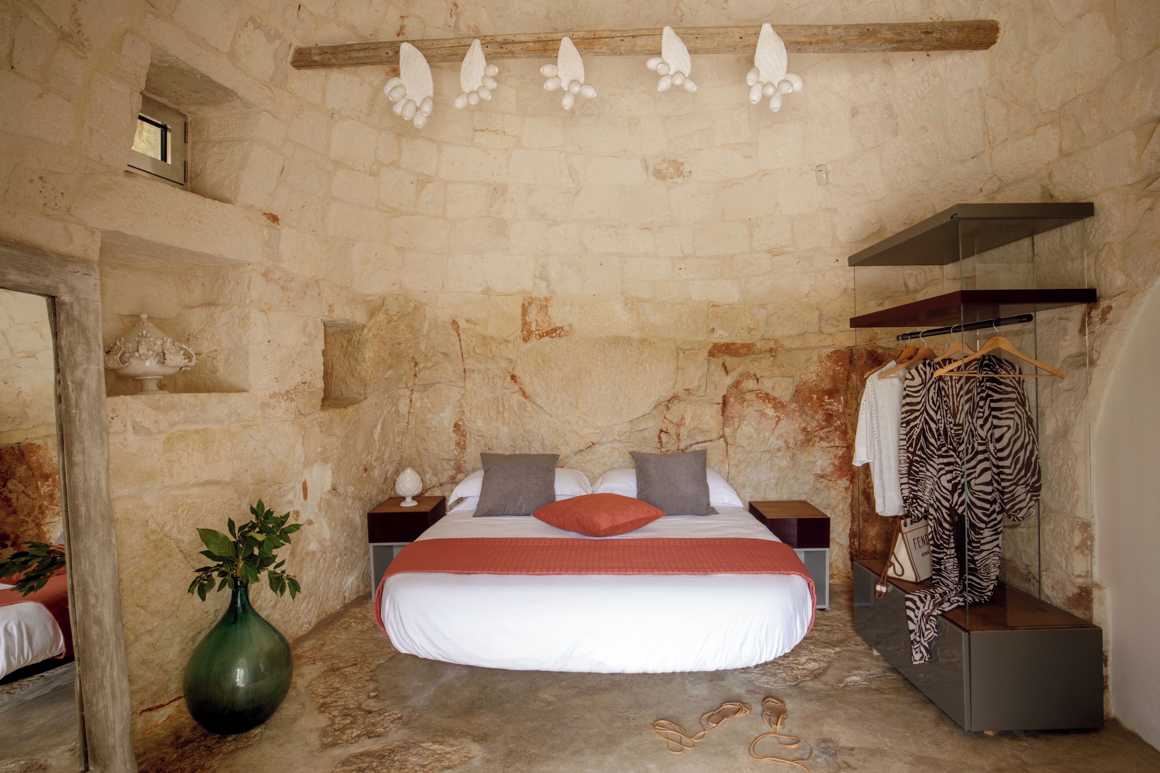 A bedroom with a stone wall, a bed with white bedding and a red pillow, a green vase, and a zebra print garment on a hanger.