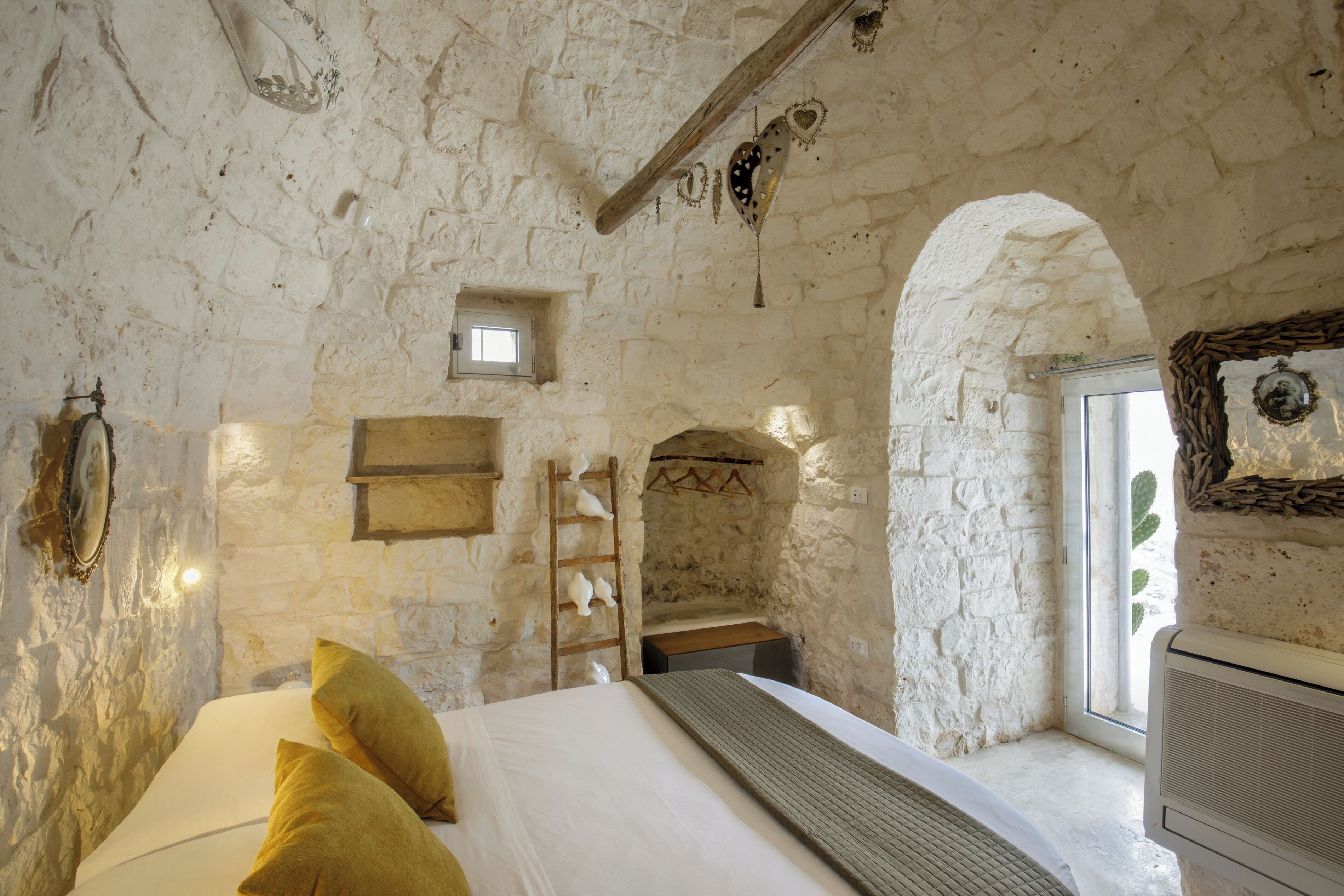 A rustic room with a stone wall, a bed with pillows, a wooden ladder, and a small window.