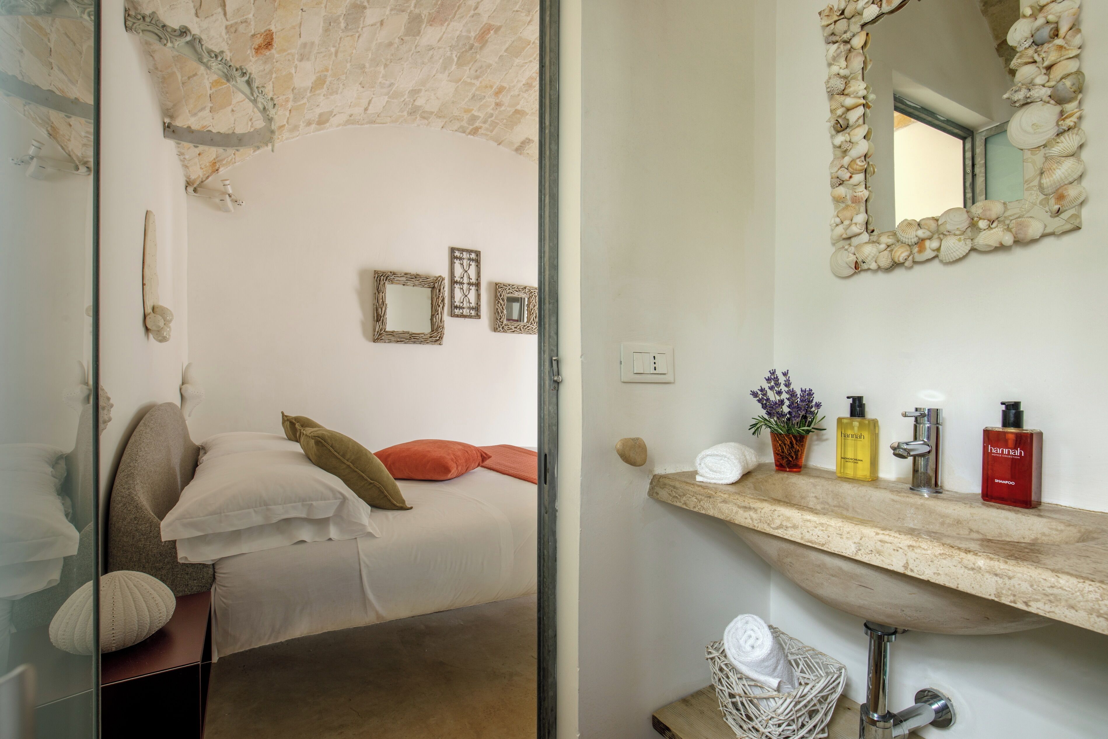 A bedroom with a bed, pillows, a bedside table, a mirror, and a bathroom with a sink, toiletries, and a stone wall.