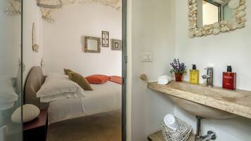 A bedroom with a bed, pillows, a bedside table, a mirror, and a bathroom with a sink, toiletries, and a stone wall.