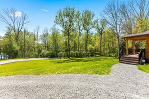 A gravel driveway leading to a wooden cabin with a porch, surrounded by a well-maintained lawn and trees. - Cozy Cabin, Sauna! Hot Tub! Filtered Lake Views! (Oakland)