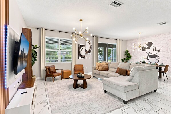 A modern living room with a flat-screen TV, leather armchairs, a sofa, a coffee table, and a Mickey Mouse mural on the wall. - Themed 9BR Villa, Pool, Game Room, Champions Gate (Four Corners)