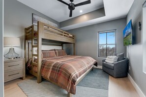A bunk bed with a plaid bedspread, a bedside table with a lamp, a gray armchair, a window with a view, and a ceiling fan. - Luxury Ski in Lodge|Indoor pool|Sleeps 21|VIEWS! (McHenry)