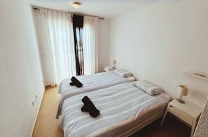 A hotel room with two beds, a window with curtains, a bedside table with a lamp, and a wall-mounted air conditioning unit. - Beautiful 2-Bedroom Apartment in Condado de Alhama Golf Resort with Pools, AC, WiFi (Los Guardianes)