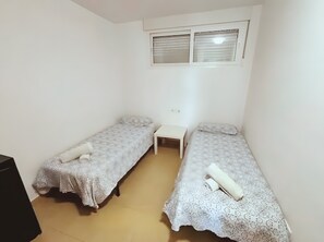 Eine kleine, einfache Unterkunft mit zwei Einzelbetten, einem Tisch und einem Fenster mit Jalousien.