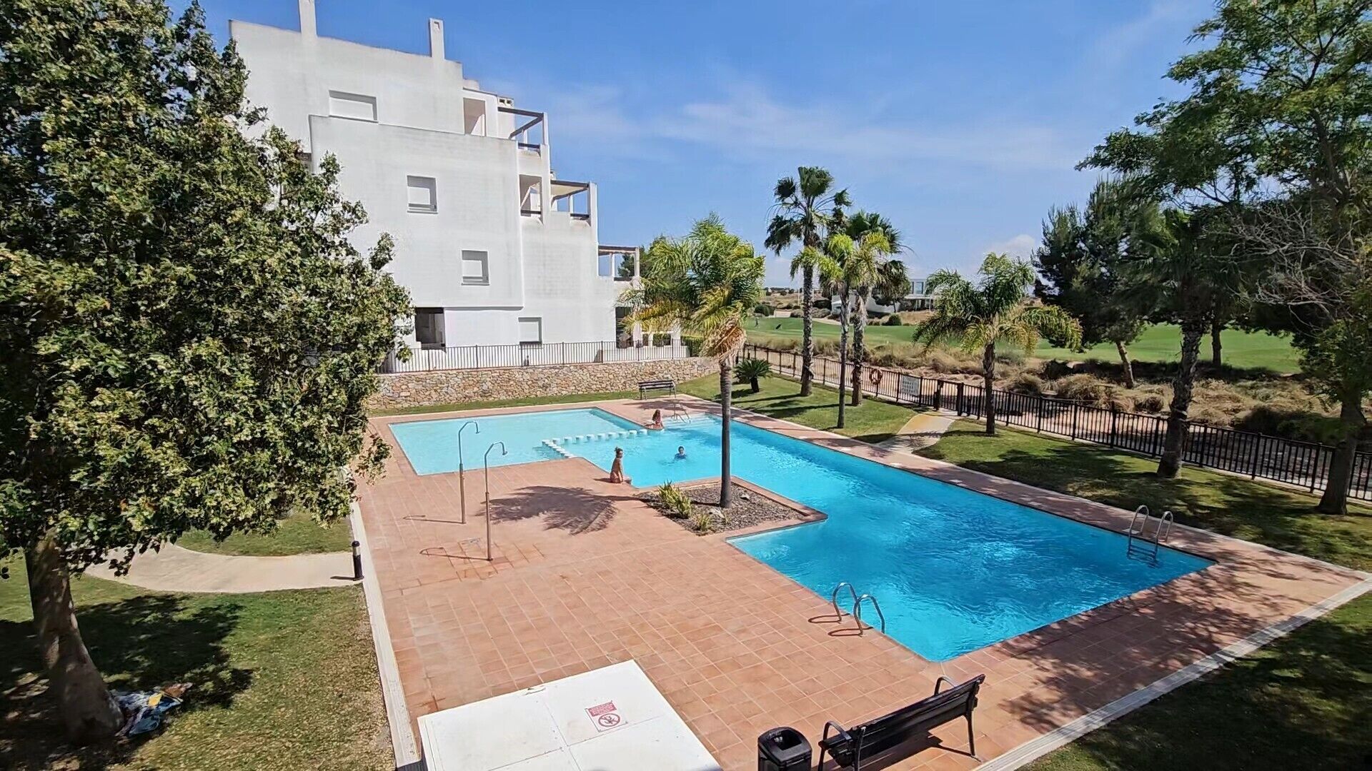 Una piscina con solana azulejada, rodeada de árboles y un edificio al fondo.