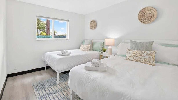 Quarto de hotel com duas camas, uma janela, um abajur e um enfeite de parede trançado.