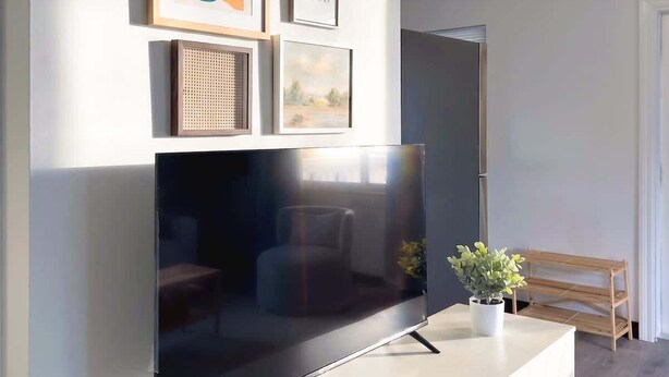 Un salon avec un téléviseur à écran plat posé sur un meuble blanc, des œuvres d’art encadrées accrochées au mur, une petite plante en pot et une télécommande sur le meuble.