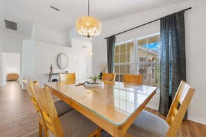 Ein Essbereich mit Tischplatte aus Glas, Holzstühlen, einer Pendelleuchte und Blick durch ein großes Fenster mit Vorhängen.