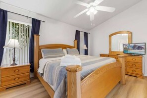 Een slaapkamer met een bed, houten hoofdeinde, nachtkastjes, een spiegel, een televisie en een plafondventilator.