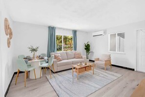 Ein modernes Wohnzimmer mit Sofa, Couchtisch und Sesseln. Ein Fenster mit Vorhängen, eine Pflanze und eine Wand-Klimaanlage sind ebenfalls zu sehen.
