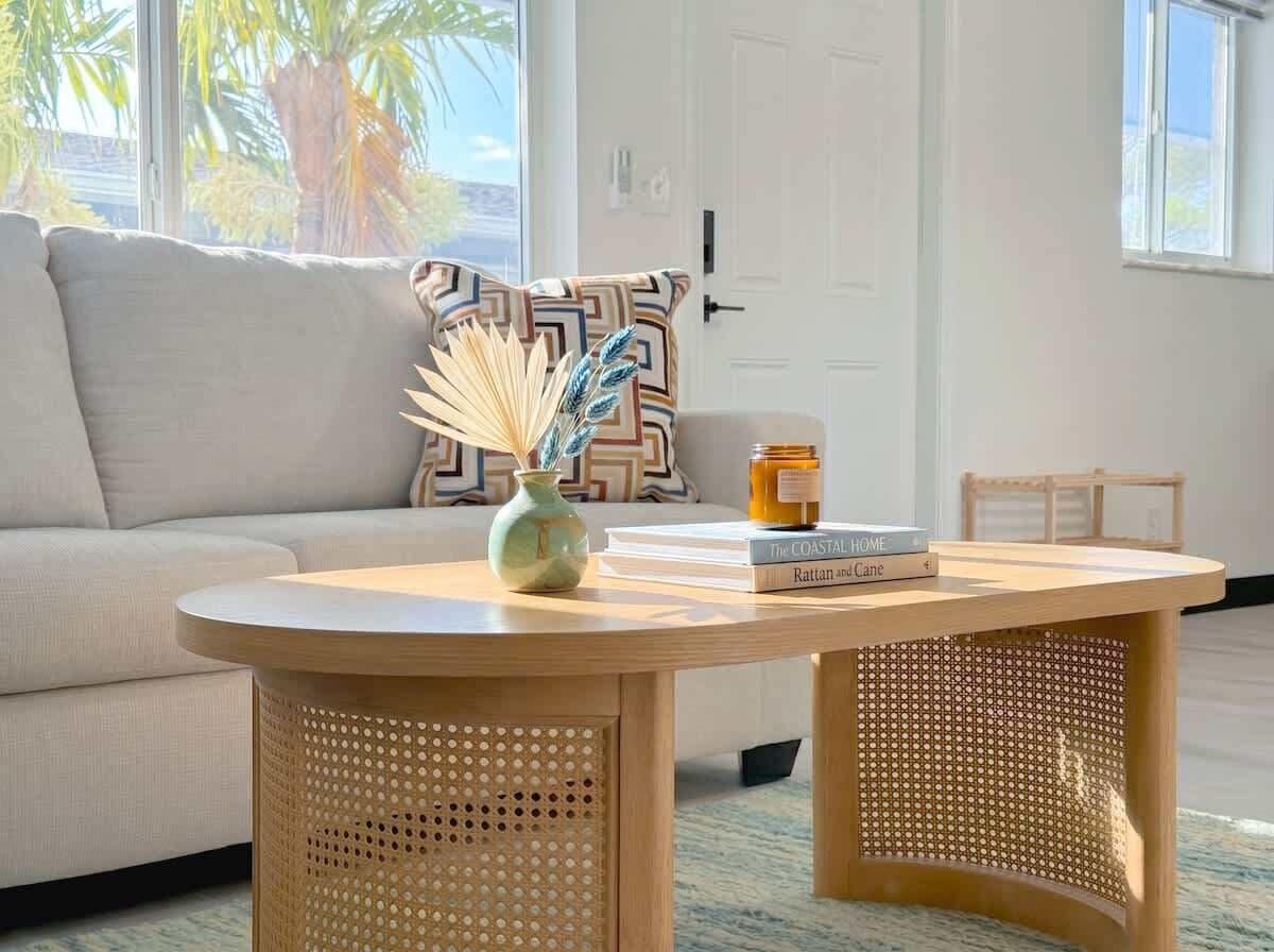 A round wooden table with a woven base, a vase with dried plants, and a stack of books.