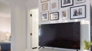 A modern living room with a large flat-screen TV mounted on a white cabinet, surrounded by framed artwork on the wall. - Inviting 2BR Near Beach, Airport & Las Olas (Fort Lauderdale)