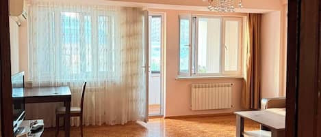 Ein geräumiger Raum mit einem gemusterten Boden, einem Esstisch mit Stühlen, einer Chandelierleuchtre und einem mit Vorhängen versehenen Fenster.