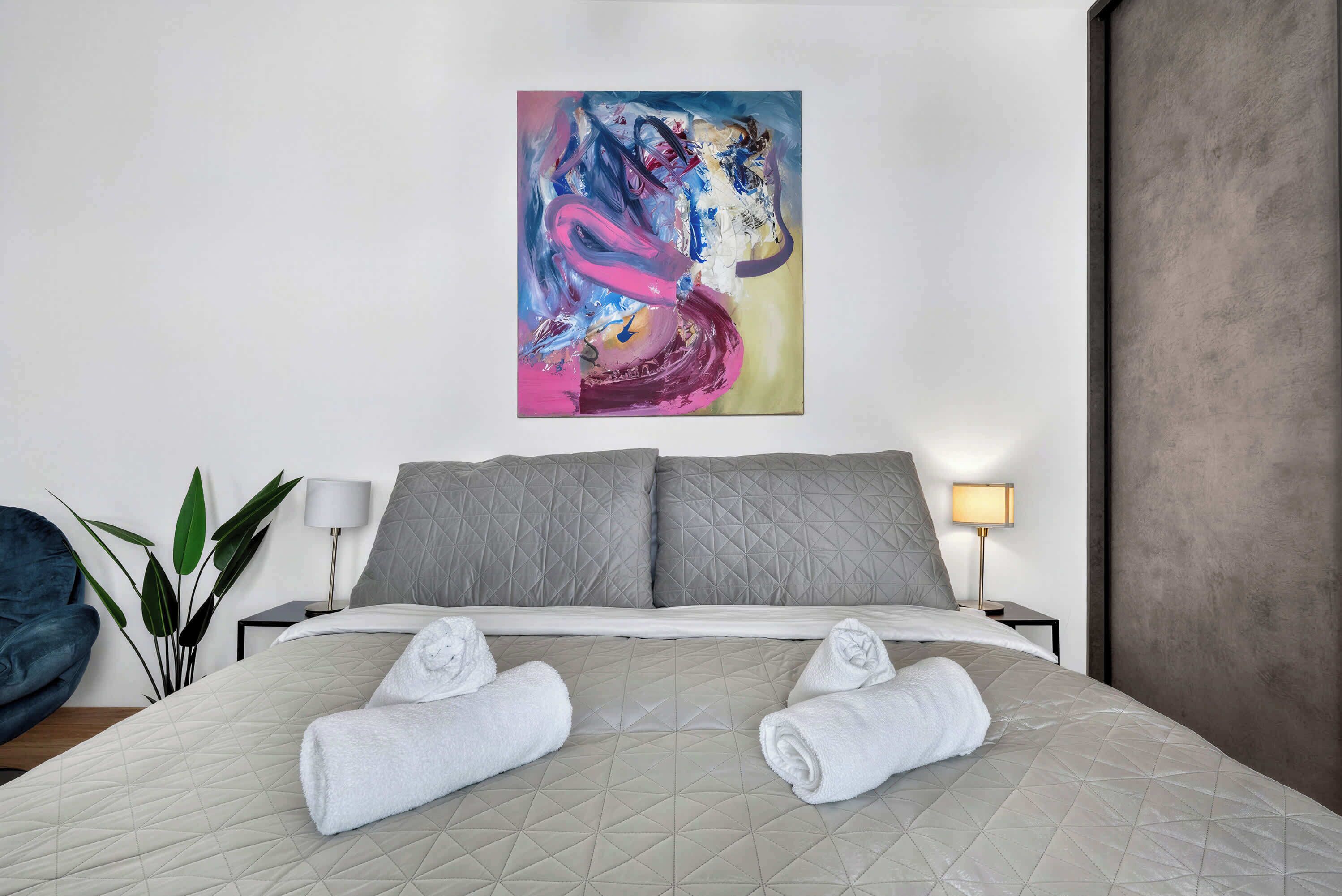 A hotel room with a bed, pillows, a painting, a lamp, and a plant.