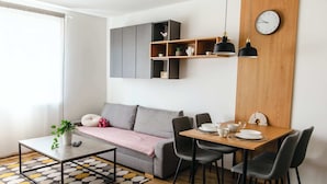 A modern living room with a sofa, coffee table, dining table, chairs, a clock, and built-in shelves. - Little Rome Loft - Vibe Apartment (Trnava)