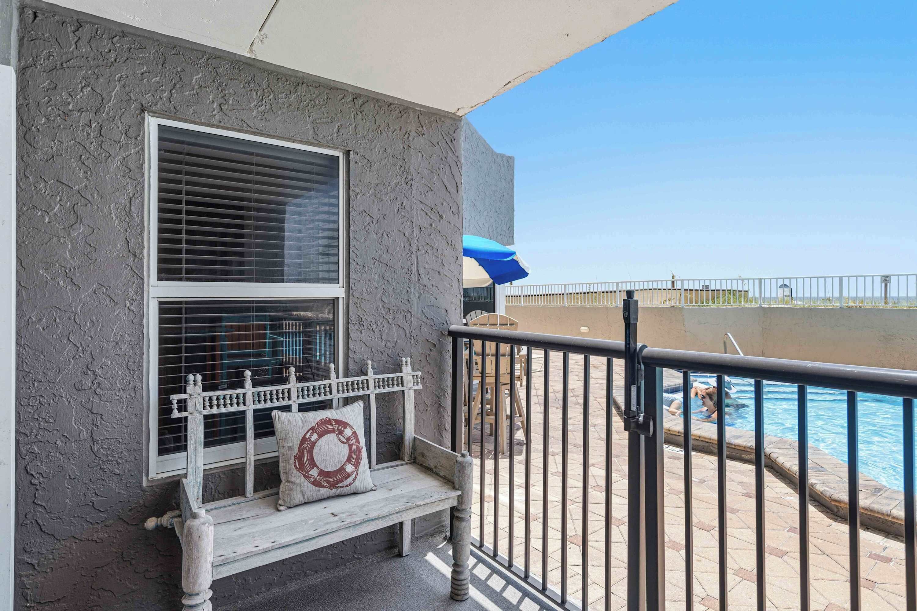 A balcony with a view of a swimming pool, a bench with a cushion, and a railing.