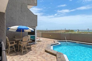 A pool area with a table and chairs, a blue and white umbrella, and a view of the beach. - Waterfront Condo with shared pool & fitness room (Fort Walton Beach)