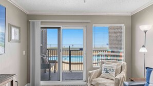 Ein Zimmer mit einer Schiebetür zum Balkon, ein Sessel mit Kissen, eine Lampe und Blick auf einen Pool.