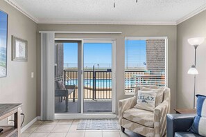A room with a sliding glass door to a balcony, a chair with a pillow, a lamp, and a view of a pool. - Waterfront Condo with shared pool & fitness room (Fort Walton Beach)
