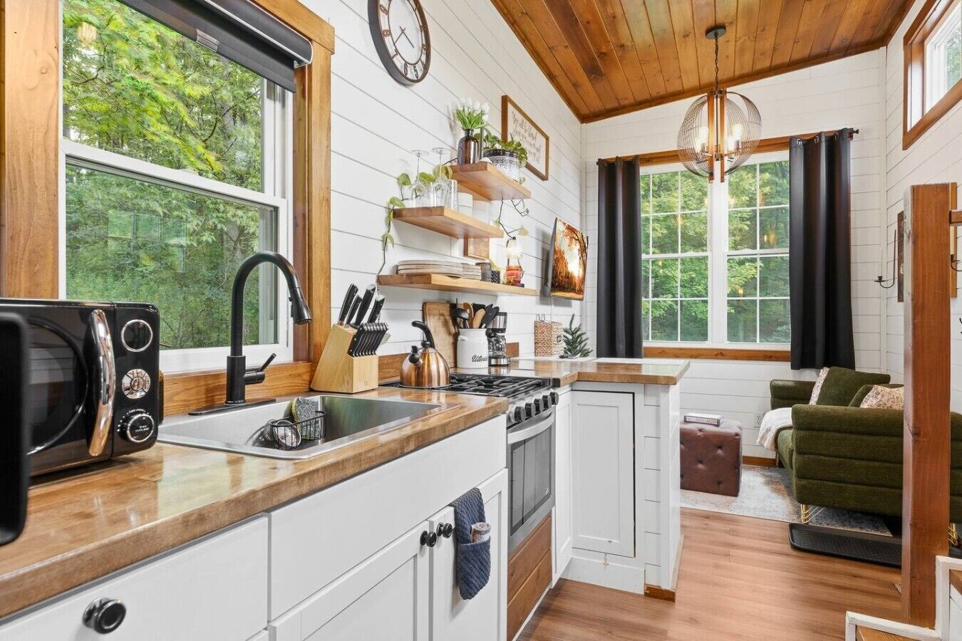 Een compacte keuken met een magnetron, spoelbak en fornuis. Boven het aanrecht zijn er houten planken, een klok aan de muur en een raam met uitzicht op groen.