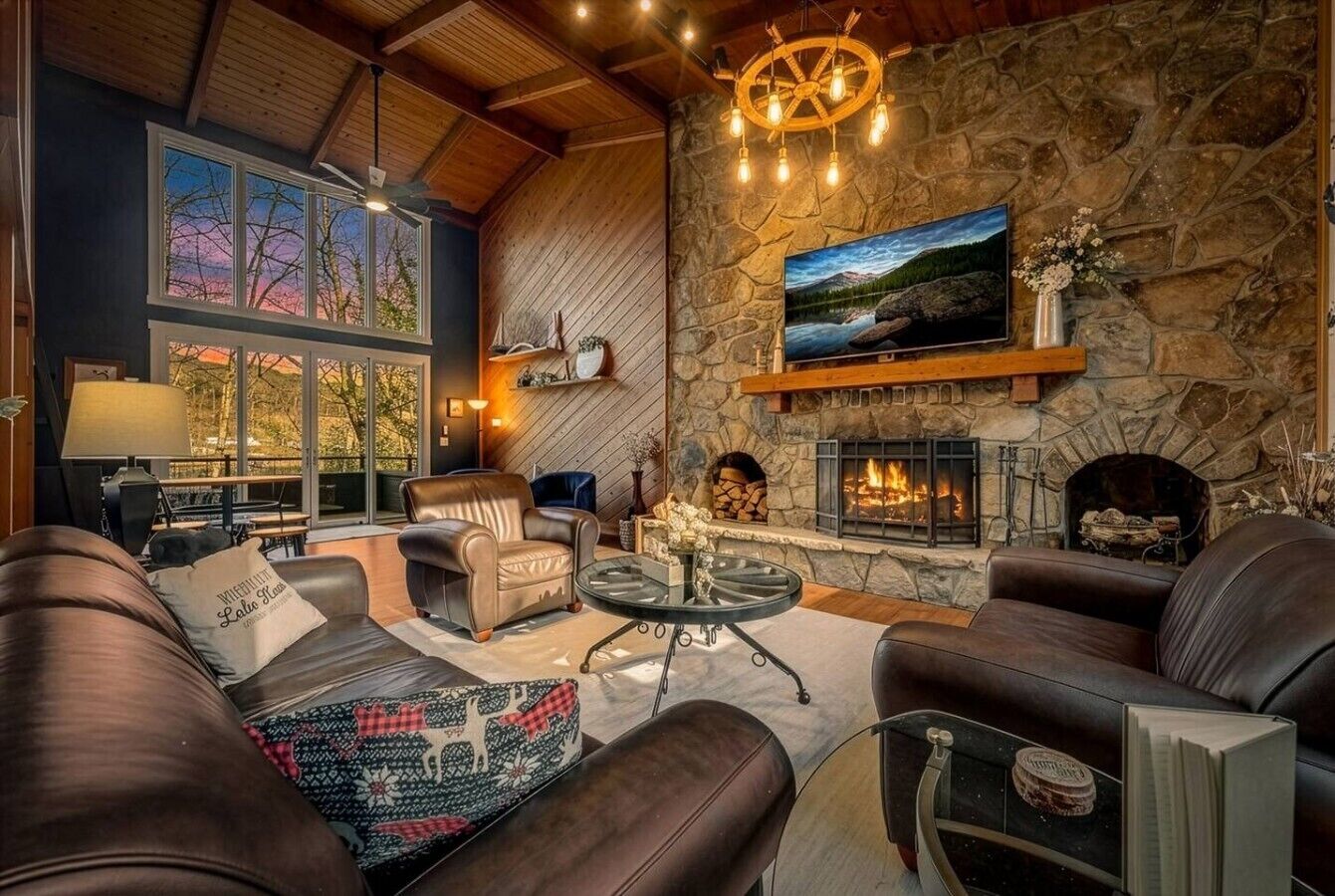 A cozy living room with leather sofas, a stone fireplace, a mounted TV, and a large window with a view of trees.