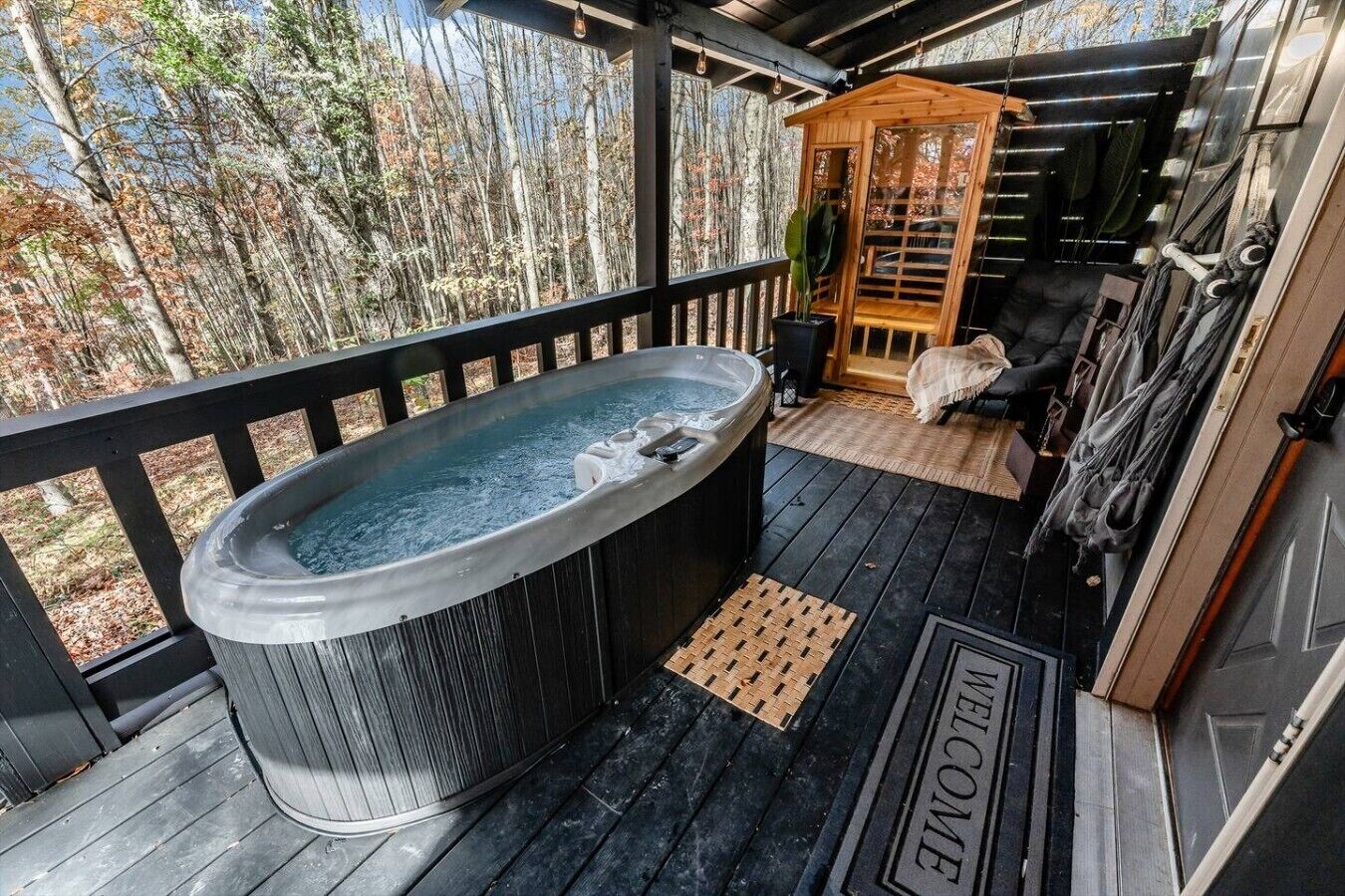 Un jacuzzi en una terraza de madera con vistas a árboles, una estructura de madera y una puerta que dice 'WELCOME'.