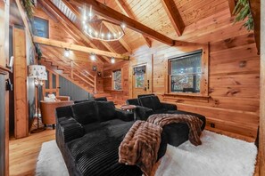 A cozy cabin interior with a staircase, a black sofa, and a chandelier.