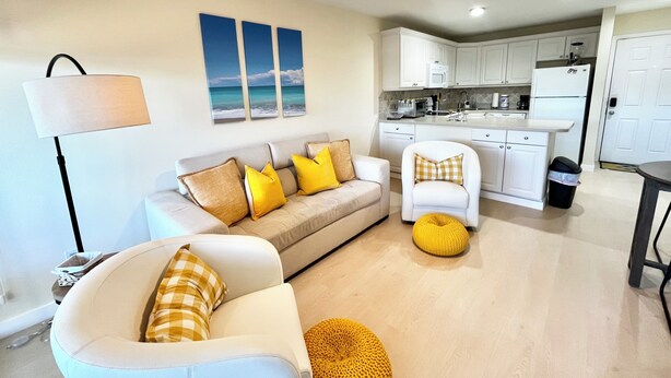 A modern living room with a sofa, armchair, and ottoman, a kitchen with white cabinets, and a ceiling fan. - OV 123 Tennis Court View - Welcome To Paradise! (Fort Pierce)