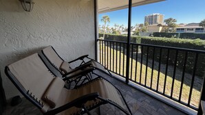 A balcony with a cushioned chair, a wall-mounted light fixture, and a view of a residential area with buildings and greenery. - OV 123 Tennis Court View - Welcome To Paradise! (Fort Pierce)