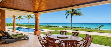 Un espace extérieur abrité comprenant une piscine, une table à manger et des chaises, avec vue sur une plage bordée de palmiers et sur l’océan.