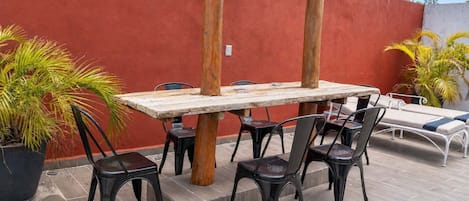 Um pátio ao ar livre com mesa e cadeiras de madeira, cercado por plantas em vasos e uma parede vermelha.