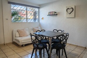 Et lille, rent værelse med en hvid sofa, et bord med stole, et vindue med udsigt til bygninger, og en væg med et skilt der siger 'BEACH' samt en hjerteformet pynt.